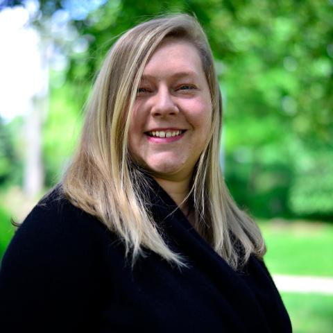 Lori has shoulder length blonde hair and wears a black sweater. She poses by some trees.