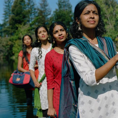 Four west Asian women wear saris in a river. They all face the same direction, toward the camera, and pose in a line.
