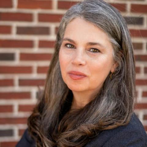 A white woman with long grayish brown hair and a dark jacket in front of a brick wall