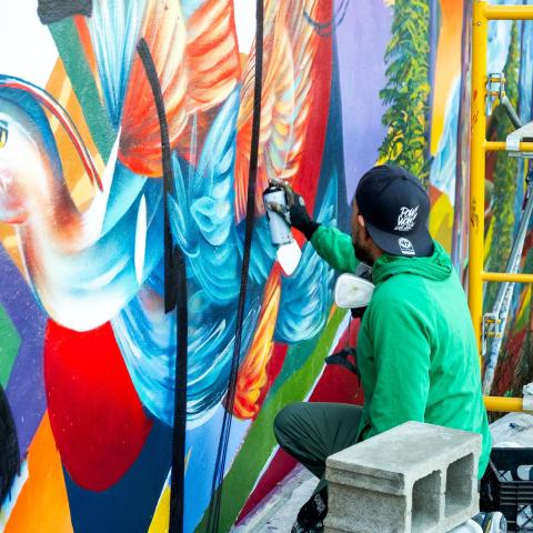 A person in a green hoodie and backwards baseball cap holds a spray paint can while working on a colorful mural