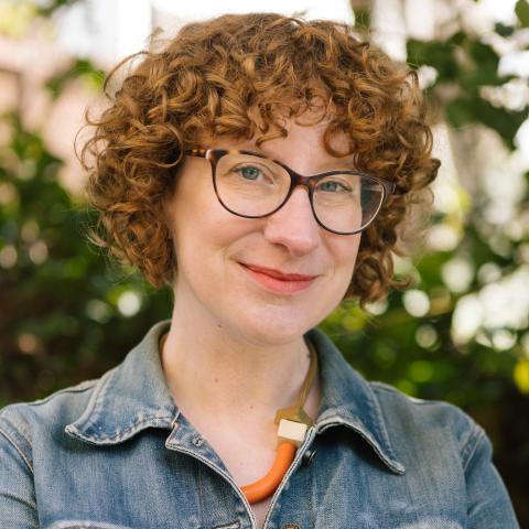 Color photograph of a white woman, early 40s, with short curly red hair, tortoiseshell glasses, and orange necklace. Pictured from chest up, standing with folded arms inside a denim jacket, against a blurry background of green foliage.