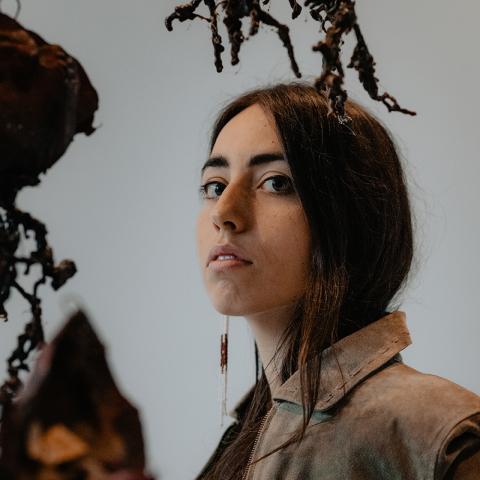 Mali has long brown hair. She wears a khaki shirt and a long earing. She poses next to earthy sculptures that hang from the ceiling.