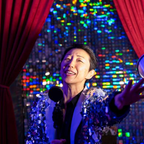 A person in a fringed jacket stands in front of a curtain and stretches out a hand.
