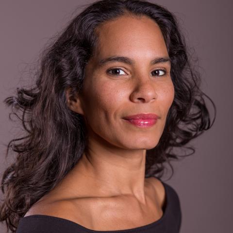 A brown skinned woman with wavy long dark hair wearing a black top with a ballet neckline.