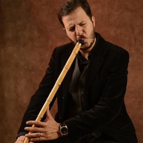 Mehmet plays a reed instrument in front of a brown background.