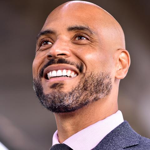 A Black man with a shaved head and short facial hair wearing a tweed jacket and tie