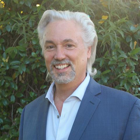 Michael is a middle-aged white man with grey hair wearing a blue blazer and open white collared shirt standing in front of a wall of white jasmine. 