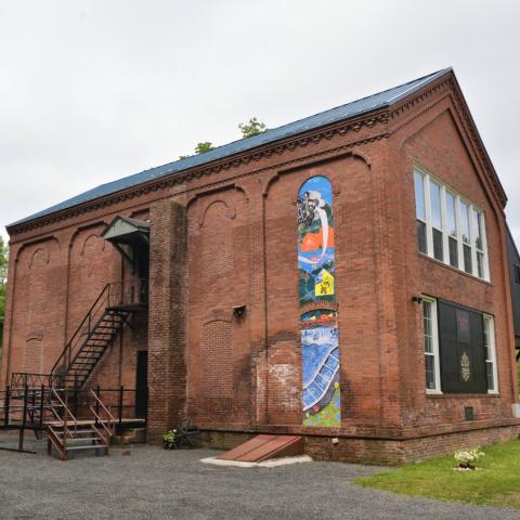 In the frame of the brick design of a small old mill, a wheatpaste mural. The design of the mural is indistinguishable from this wide shot showing the whole building.