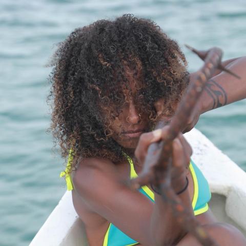 A Black woman, in a bikini and with shoulder length curly hair, aims an anchor or sling shot or some kind of metal piece at the camera.