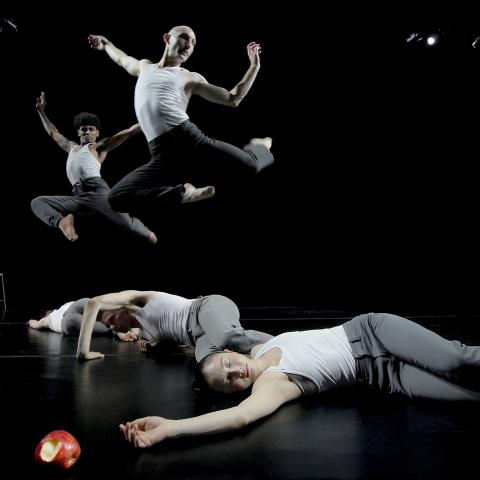 Two male dancers leap behind three female dancers who are unconscious next to an apple with a bite taken out of it.