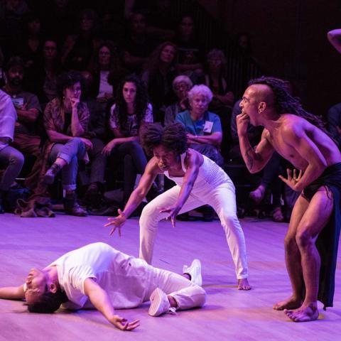 A man dressed in Native American garb pretends to vomit on two women who are dancing in white and look to be exorcising a demon from one of the women
