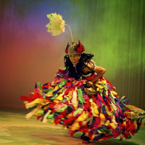 A woman in a multicolored gown dances in a space that reflects multicolored light like a rainbow