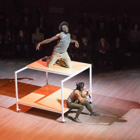 A man on top of a large table-like structure dances and a woman sits beneath it speaking to the audience.