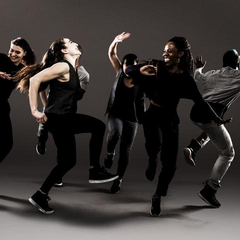 A group of break dancers dance. A couple women in the foreground appear to share a joke.