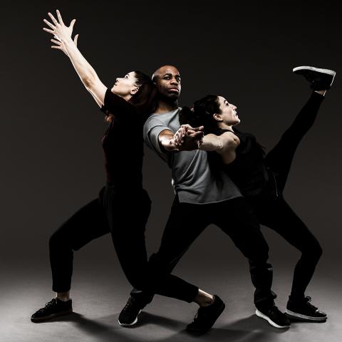 Two women lean against a male dancer. One female extends her arm and the other kicks her leg almost up to her face.