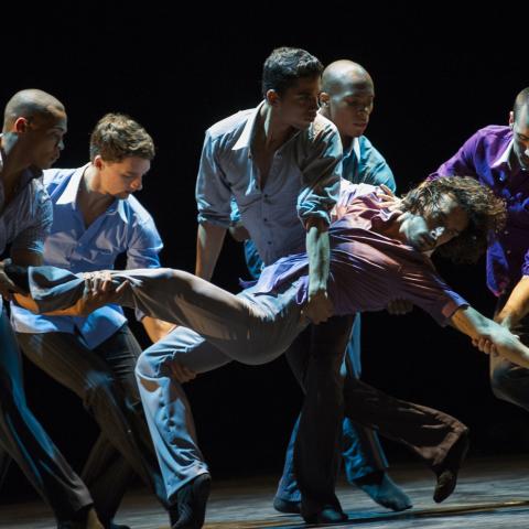 Five dancers on a dimly lit stage support a sixth dancer