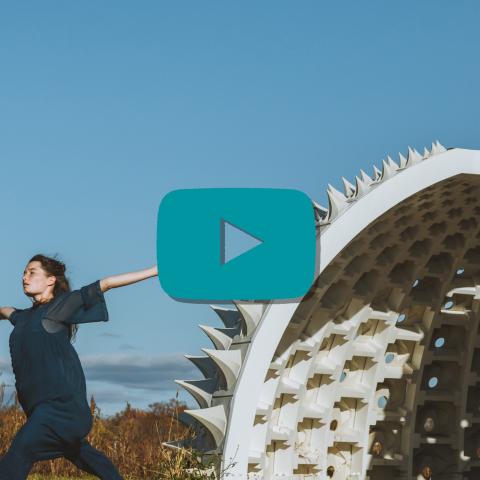 A teal play button over a light-skinned woman, outside and in all navy, holds her arms out; she stands in front of a structure that's white and has holes and spikes in it.