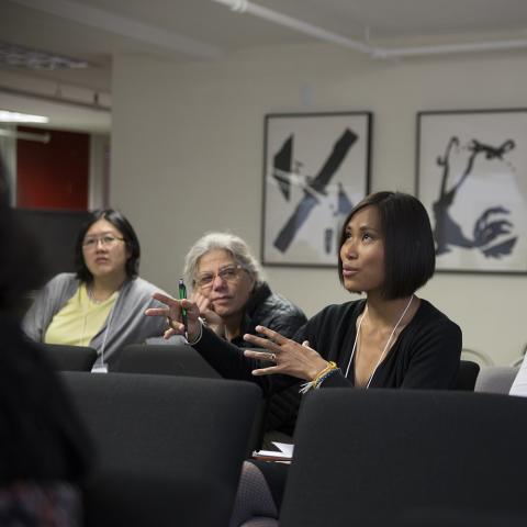 A woman speaks using hand gestures while sitting in a crowd of women