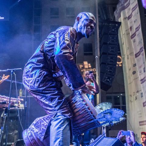 Outside, a Black man plays the drum with his hands and behind him there are other instrumentalists.