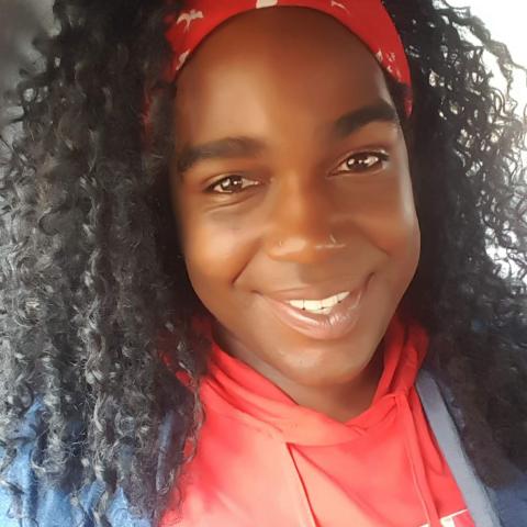 Natalie is a Black womxn with curly hair. They wear a red bandana as a headfband and a matching red sweatshirt.