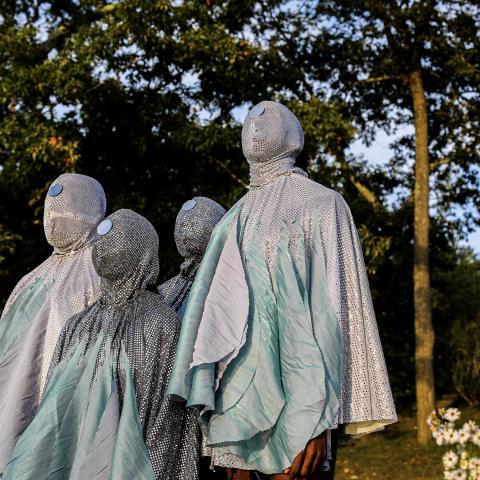 Four people wear bejeweled costumes that cover their faces. They pose plainly in front of trees.