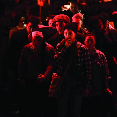 A young person in a crowd of people that appear to be in a riot holds something up to their mouth