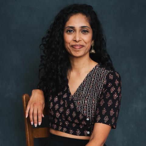 Nadhi is a brown woman with long black hair. She sits on a wooden chair in front of a grey/green backdrop. 
