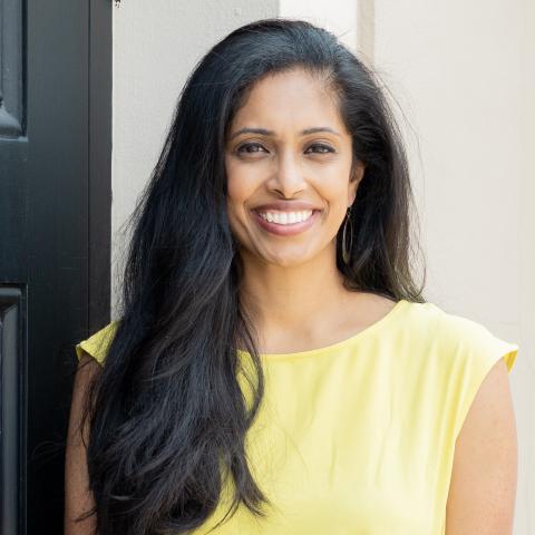 Nadhi is a brown woman with long black hair. She wears yellow and smiles brightly.