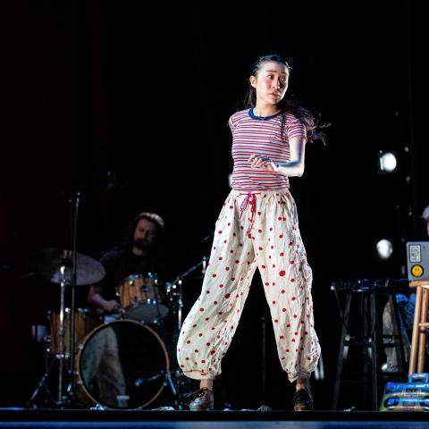 An Asian woman, in baggy pants, walks across a stage.