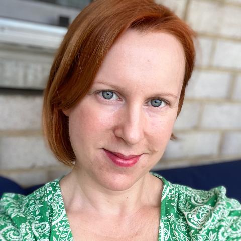 Sara Nash is a white woman with shoulder length red hair. She is outside, on a porch, and in a green paisley blouse.
