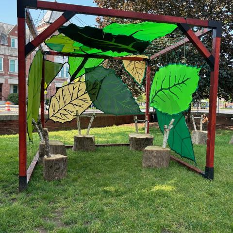 A structure with a wooden frame and with 3 walls and a ceiling defined by large leaves of shades of green and tan. Inside the space are 5 seats made of tree stumps with birch branch seat backs. The structure is on a grassy, shady area outside of a brick town building on a sunny day.