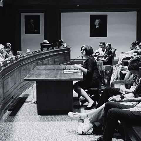 In a courtroom, a woman sits behind a table and before a group of folks.