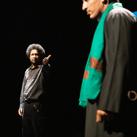 On a stage and under spotlights, a Black man, in all black, holds his hand out to a brown man in a green scarf and black robe.