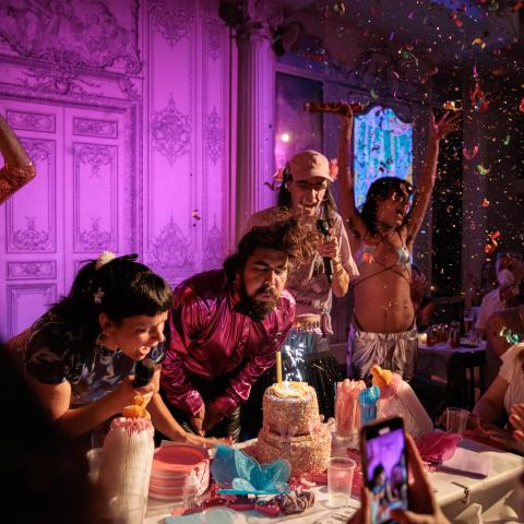 A lively birthday celebration with a group of people gathered around a table at Bowery Poetry Club. In the center, a man and a woman are blowing out candles on a colorful cake while others cheer and celebrate. The scene is vibrant, with confetti flying in the air and party decorations visible, including pink and blue napkins, plates, and utensils. The background features an ornate, pink-lit wall with detailed patterns, adding to the festive atmosphere. Liz and Bow are holding microphones.