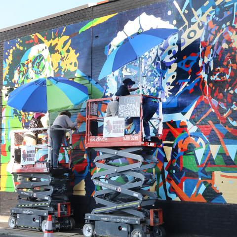 Outside, two folks, on risers, paint a mural on the side of a brick building.