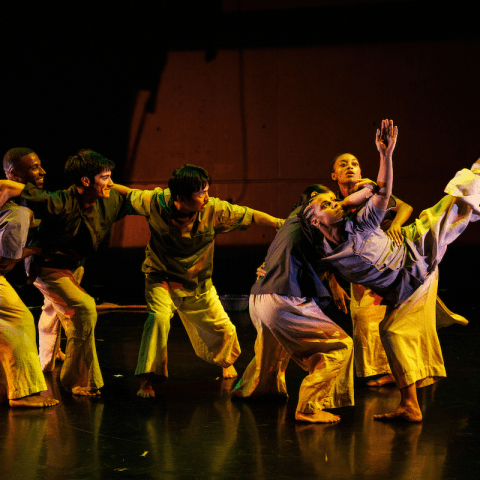 A group of six performers are on a stage with their arms slung over each others shoulders. The one on the far right raises a leg and arm towards the sky.