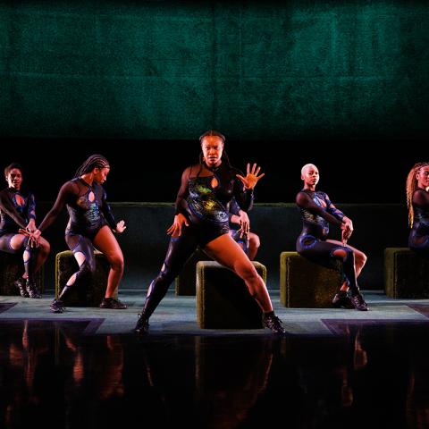 On a dark stage, five women of color either sit on blocks or stand and face the audience. They all wear asymetrical body suits.