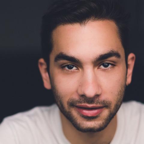 a head shot of a young man with a beard wearing a white shirt. The backdrop is all black.