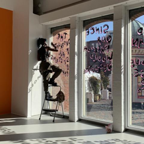 A Black womxn paints text on glass windows. In an orange frame, another image with "How Will You Create Public Life?" painted on yellow.