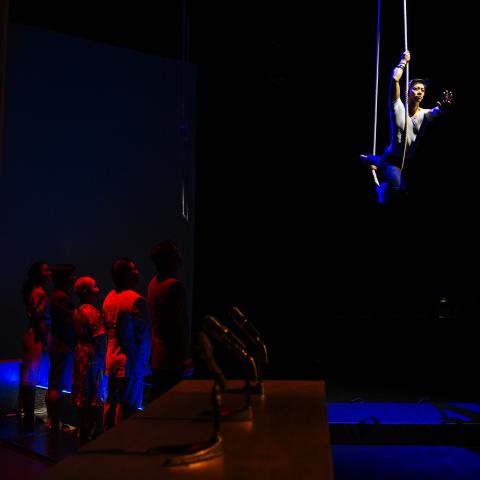 A performer swings from a rope, while other performers stand below on a stage in red light.