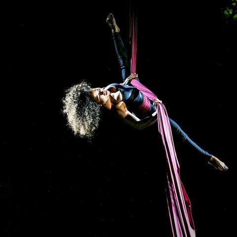 In a black void, a black woman with an afro gives an aerial performance with fabric.