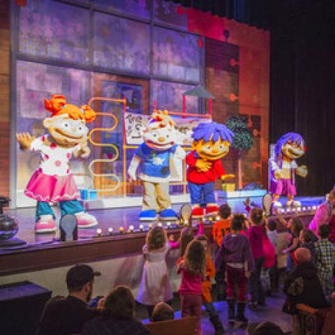 Actors dressed in full body costumes, with oversized heads perform on a stage decorated in bright colors.