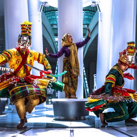 In a mall, with pillars and round, backless benches, a woman poses on a bench and two folks in ornate red, yellow, and green costumes twirl.