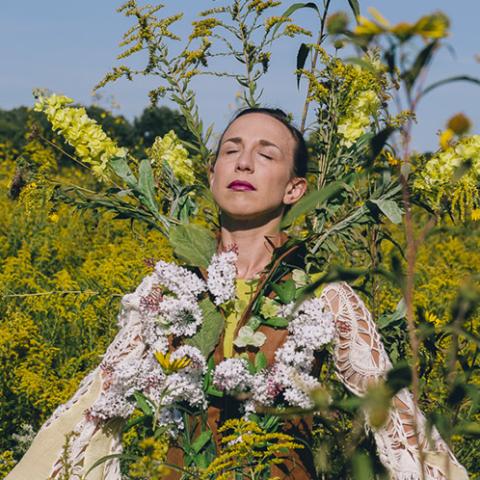 A white woman stands in field, intertwined with tall flowers.