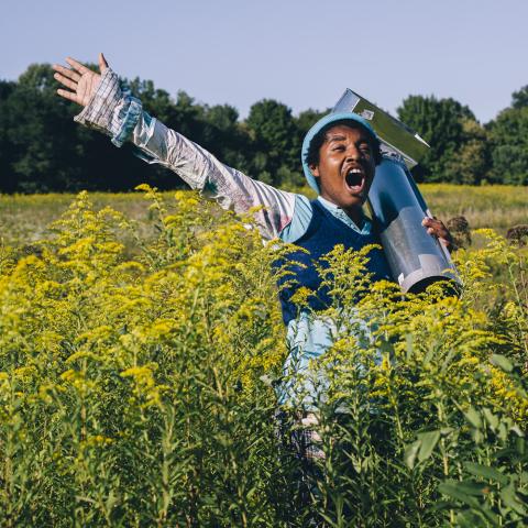 A young Black person carries something shiny in a field of flowers and screams.