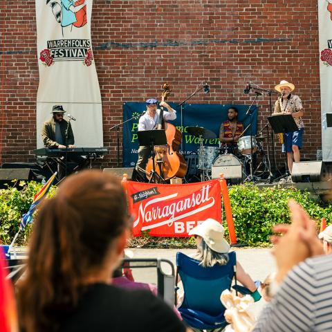 On an outdoor stage and in front of a brick wall, Black and white folks play jazz instruments while blurry folks watch in the foreground.