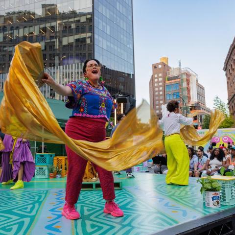 On an outdoor stage in a city, performers hold fabric and sing admist brightly colored set pieces.