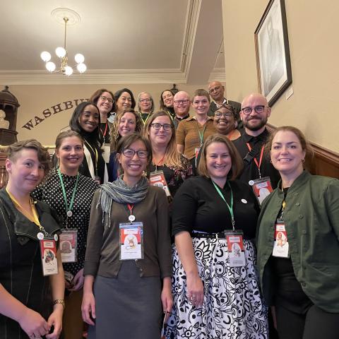 Inside, on stairs, about twenty people pose together with name tags.