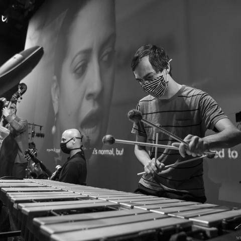 A man plays the symbols in front of a projection of a woman speaking.