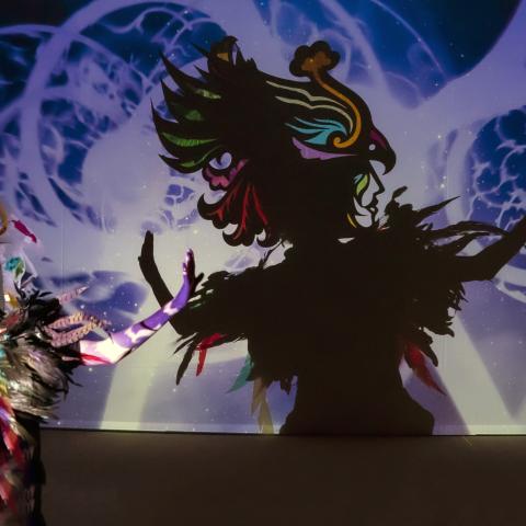 On a stage, a person in feathers and a headpiece stands in front of a spotlight. On the backdrop behind them, an animated shadow and clearer representation of the character appears.